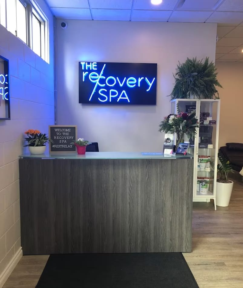 The reception area of The Recovery Spa in Kelowna, featuring a wooden counter, potted plants, and a neon sign that reads The Recovery Spa.