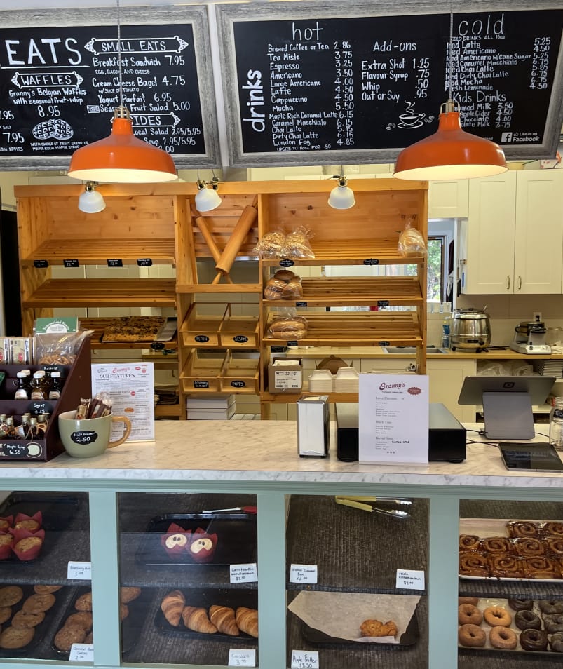 A cozy bakery in Summerland featuring various pastries and drinks, with a blackboard menu listing sandwiches, small eats, and beverages above the counter.