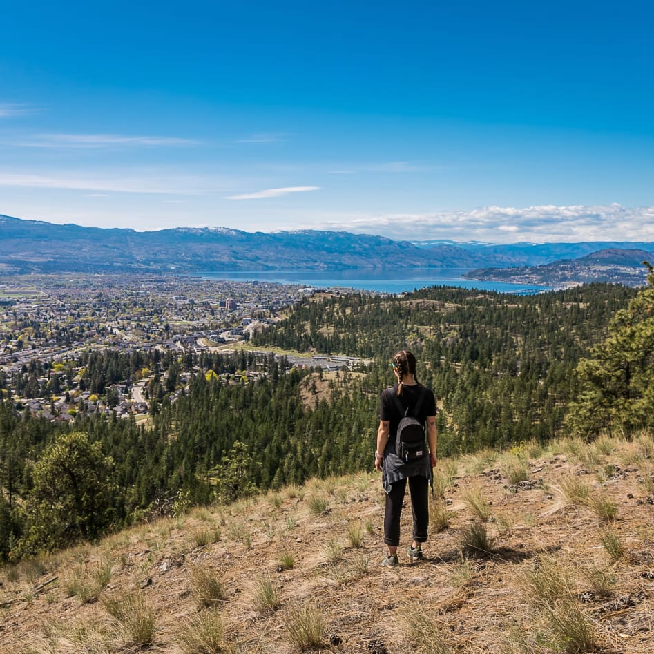 A person with a backpack overlooking the cityscape and mountainous landscape of Kelowna under a clear blue sky.