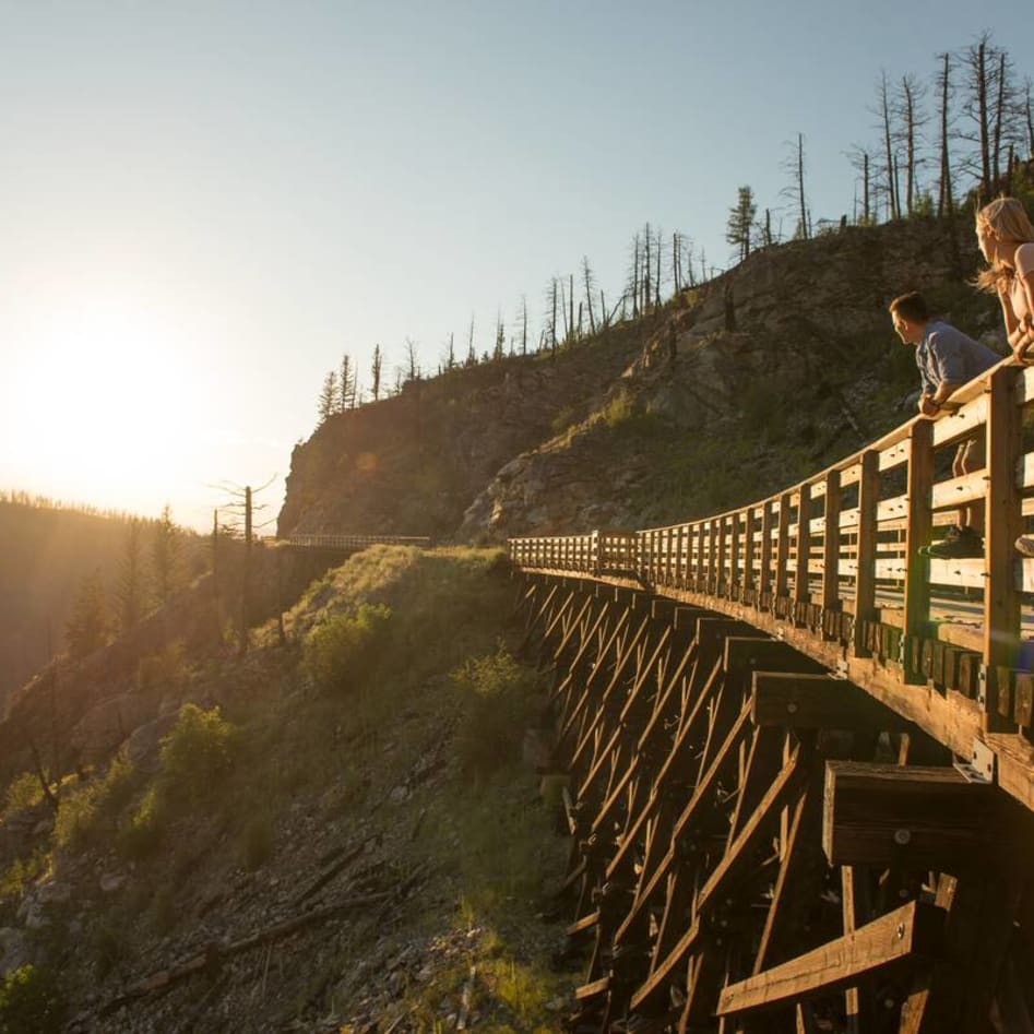 Two people enjoy the view from a wooden trestle bridge at sunset in a mountainous area.