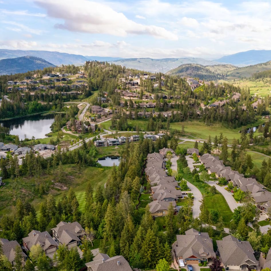Aerial view of a residential area in Vernon surrounded by green hills, trees, and small bodies of water under a partly cloudy sky.