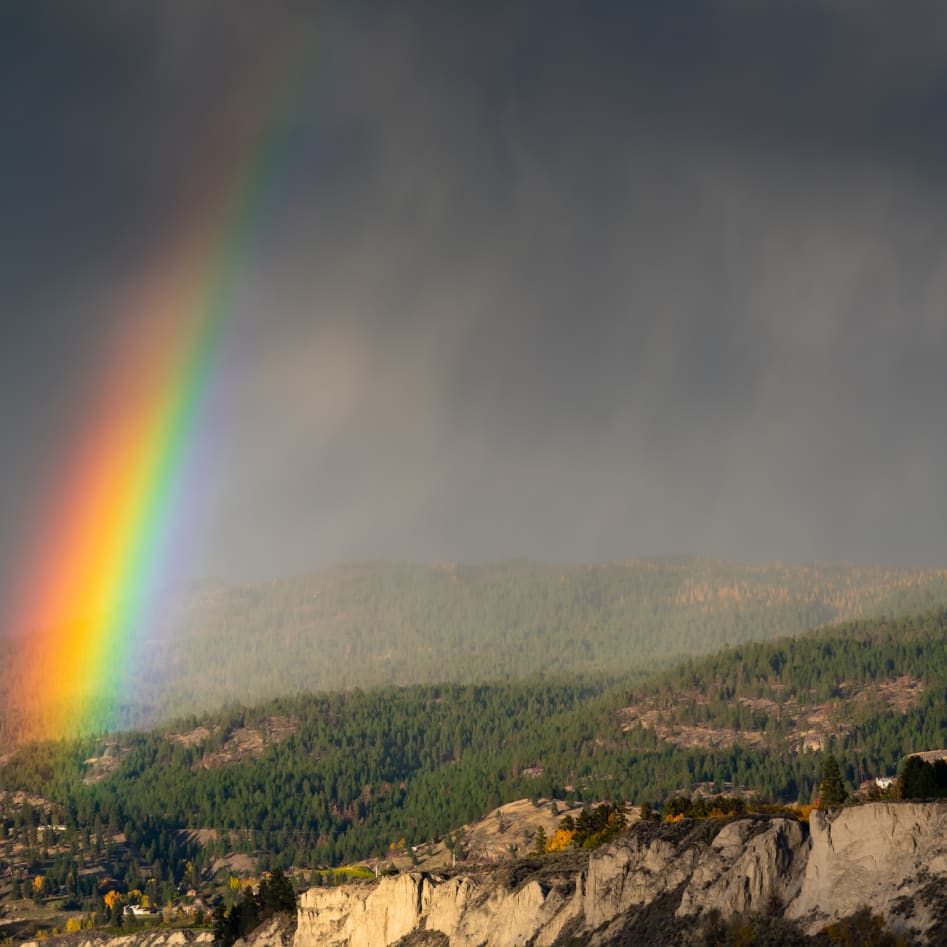 A vibrant rainbow over the lush green hills of Penticton under a dramatic, cloudy sky.