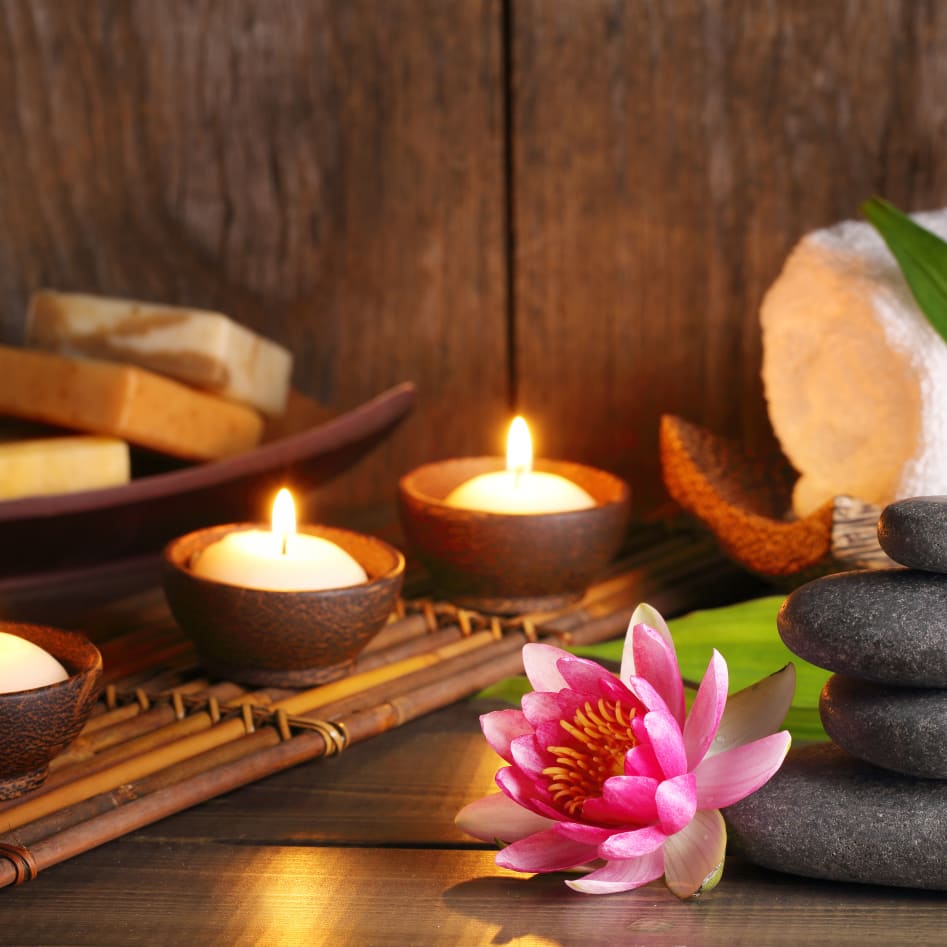 Spa scene with lit candles, stacked stones, a pink flower, soap bars, and a rolled white towel on a wooden surface.