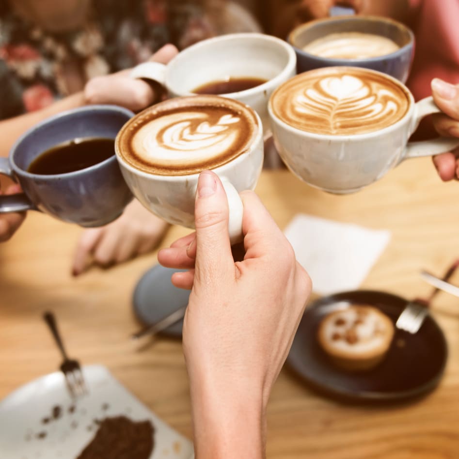 A group of people toasting with coffee mugs above a table with food and pastries.