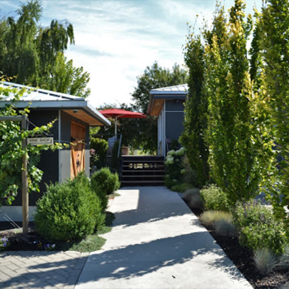 A charming outdoor view of 8th Generation Vineyard in Summerland, featuring a walkway lined with lush greenery and a sign reading "Wine Shop."