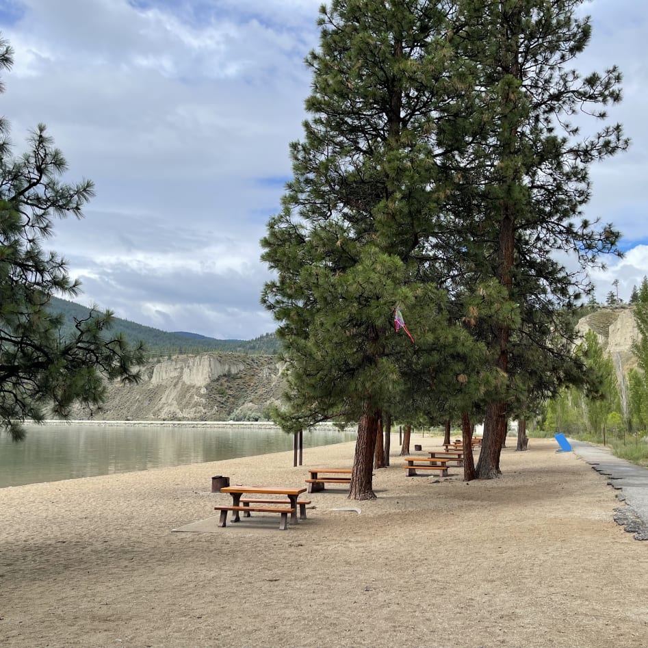 A peaceful lakeside beach at Summerland, with picnic tables and tall trees under a cloudy sky, mountains in the background.