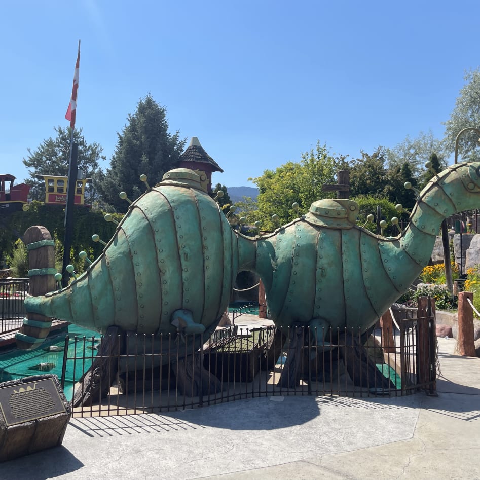 Large green dinosaur statue with bolts and knobs in an outdoor amusement park setting on a sunny day, Penticton.