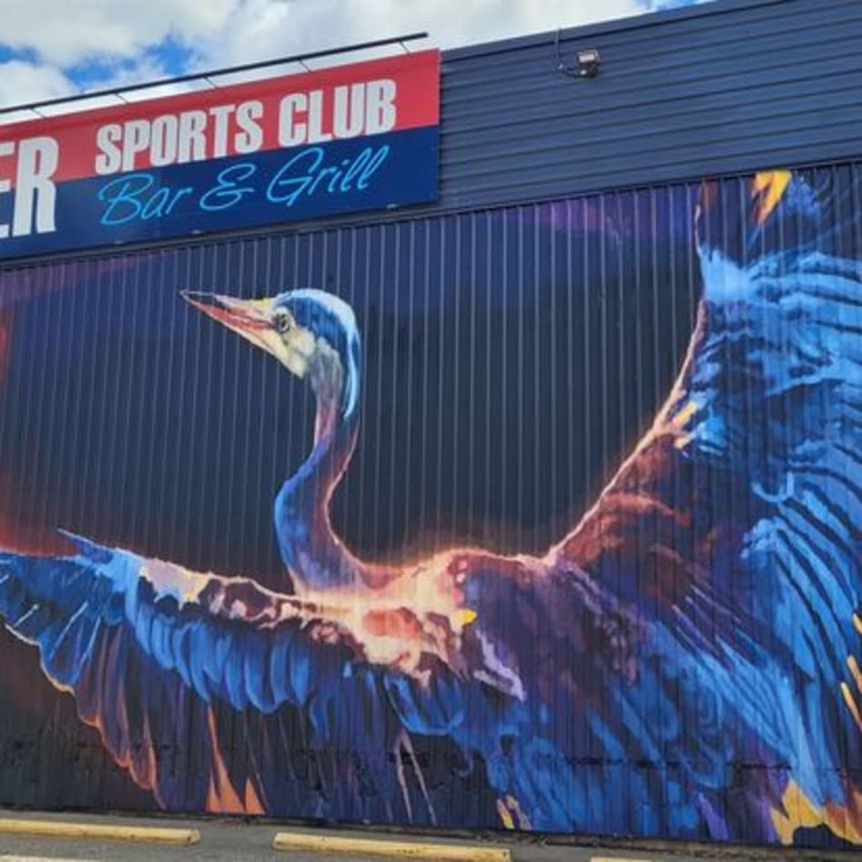 A vibrant mural of a large bird with outstretched wings adorns the side of a Vernon sports club bar and grill building.