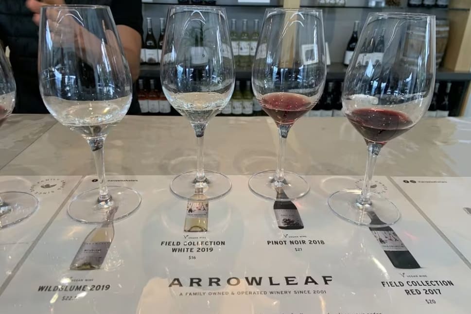 Four wine glasses sitting on top of a white counter next to a bottle of wine and a glass of wine, featuring Wildblume 2019, Field Collection White 2019, Pinot Noir 2018, and Field Collection Red 2017 by Arrowleaf Cellars