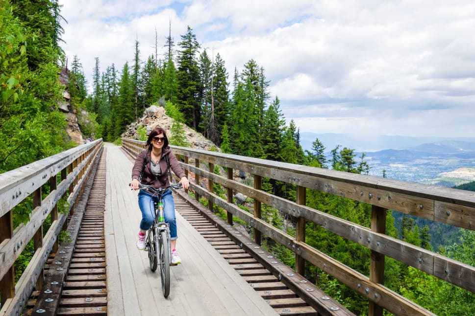 A person rides a bicycle on a wooden bridge surrounded by lush green trees and mountains in Kelowna.