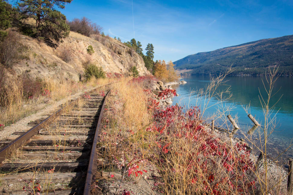 An abandoned railway track winds along the grassy edge of a lake with mountains in the background in Kelowna, under a clear blue sky.