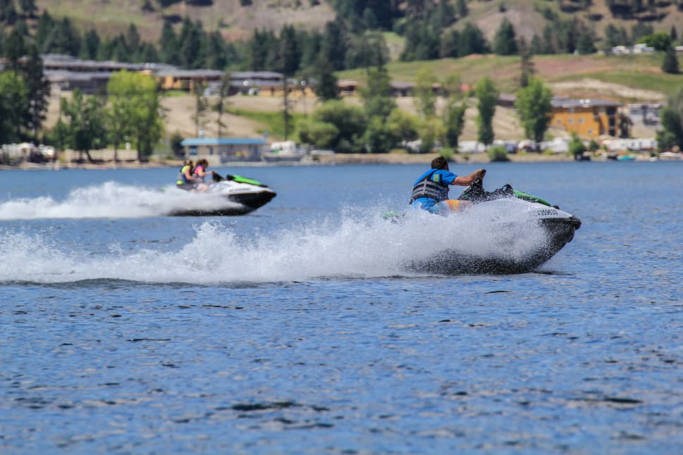 Two people riding jet skis on a lake with trees and buildings in the background in Kelowna.