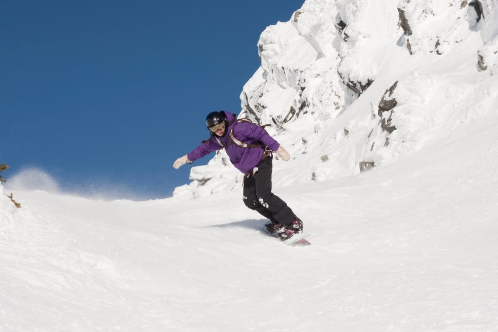 A person snowboarding down a snowy slope with rocky cliffs in Kelowna under a clear blue sky.