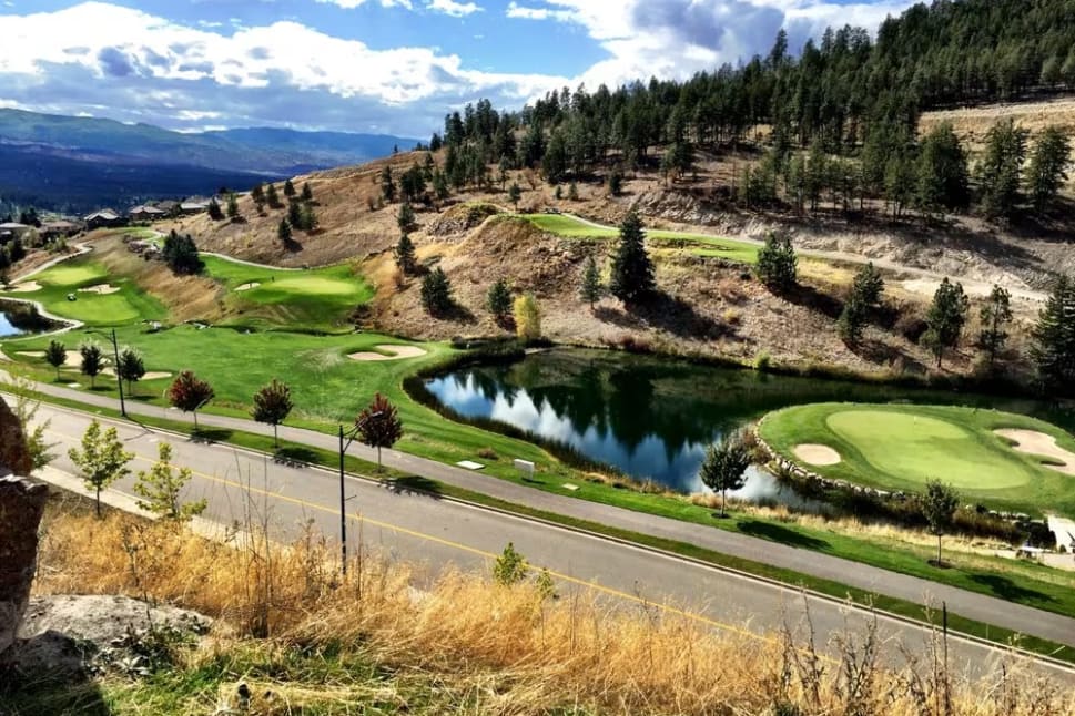A scenic view of Black Mountain Golf Club in Kelowna featuring lush greens, water hazards, and a backdrop of rugged hills and trees under a partly cloudy sky.