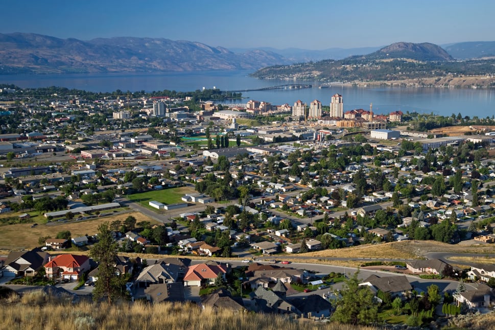 Aerial view of Kelowna showing residential houses in the foreground, the cityscape, and a large lake with mountains in the background.