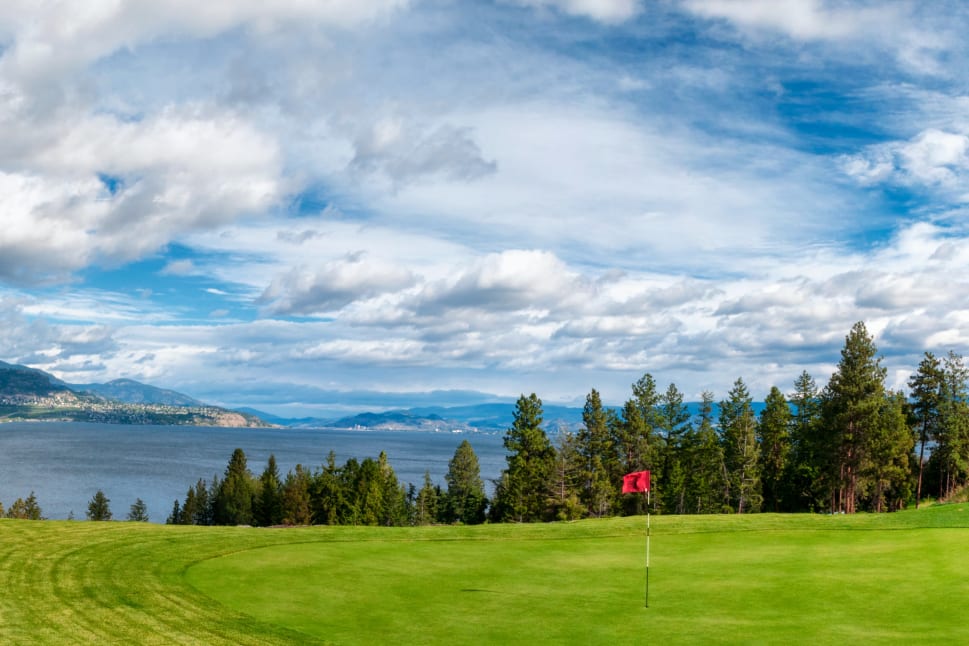 A golf course with a red flag on the green, overlooking a lake and surrounded by trees, mountains, and a partly cloudy sky in Kelowna.