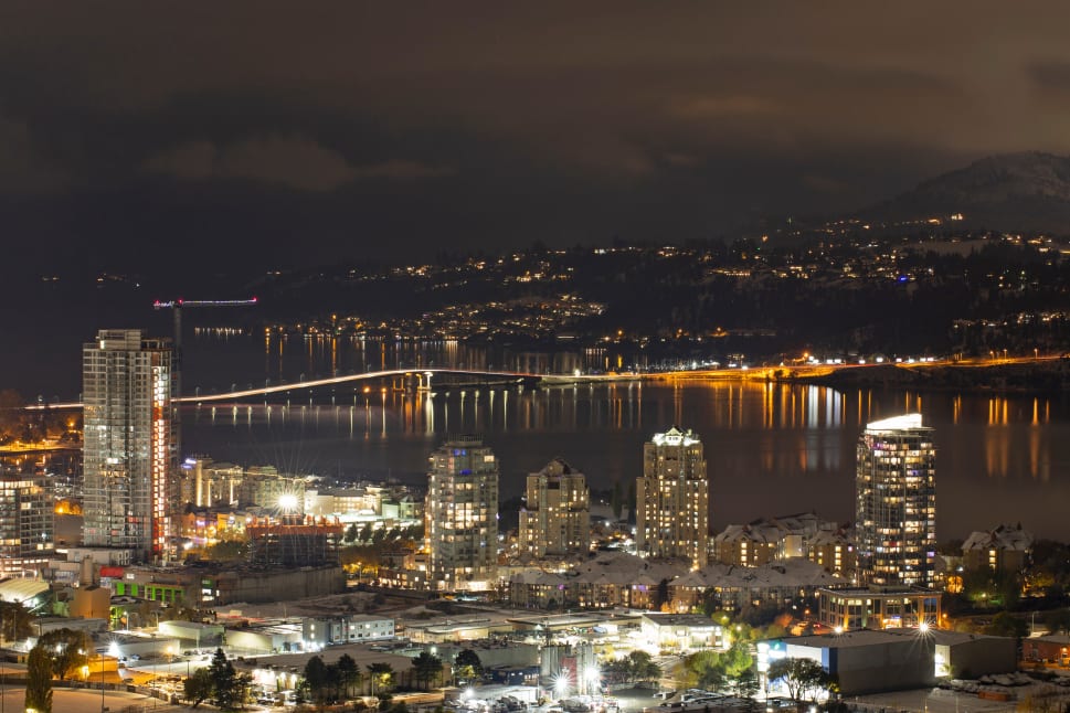 Nighttime cityscape of Kelowna with lit-up buildings, a bridge, and a dark mountainous background.