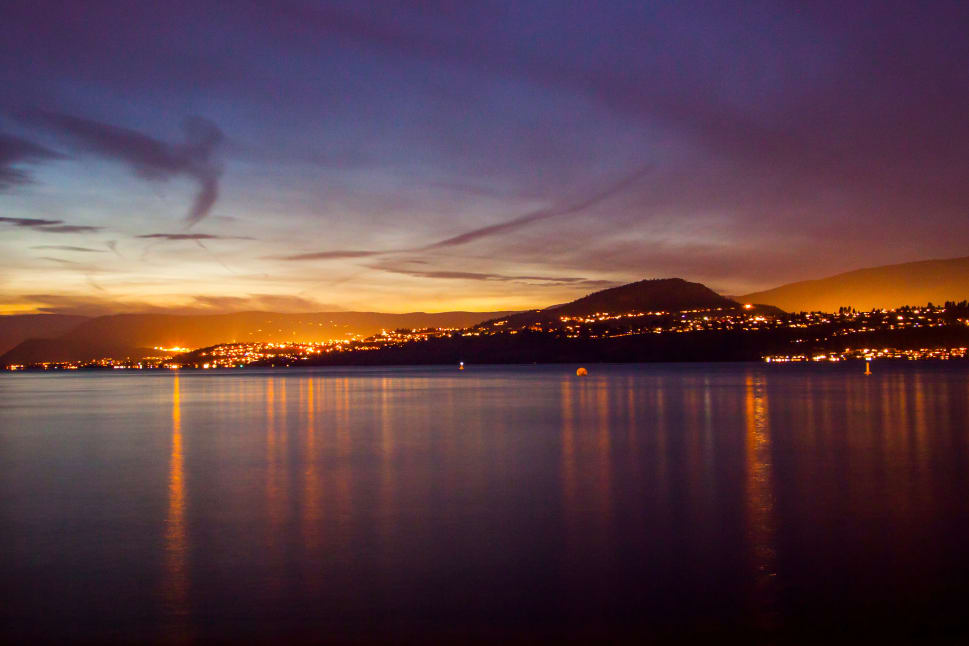 A serene dusk view of Kelowna with city lights reflecting on the calm waters of Okanagan Lake under a twilight sky.