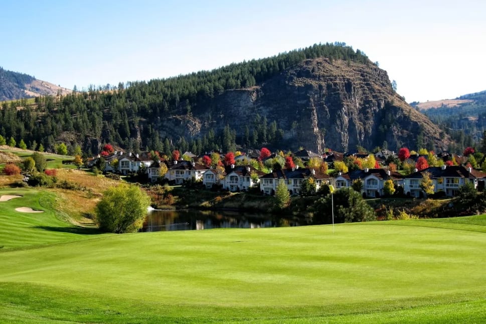 A scenic view of Gallagher's Canyon Pinnacle Course in Kelowna, featuring green fairways, a picturesque pond, homes nestled among vibrant autumn trees, and a rocky hill covered with pine trees in the background.