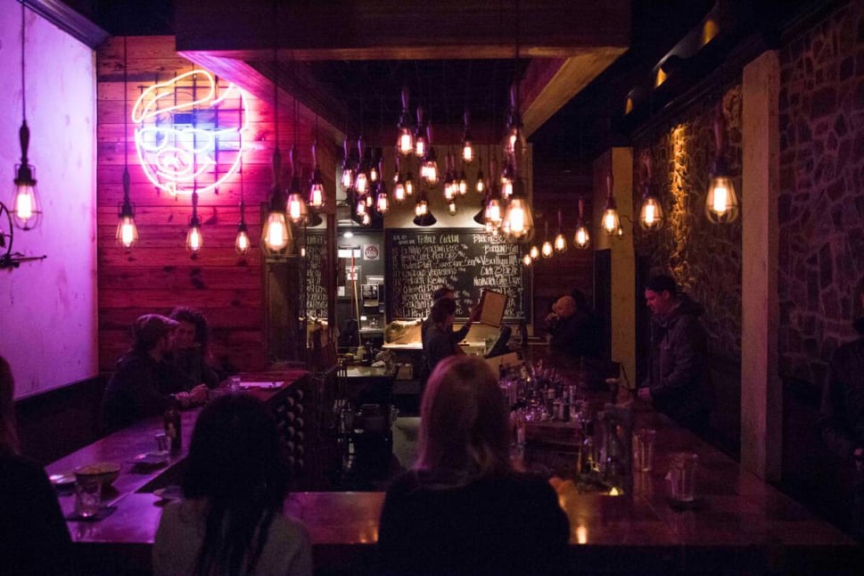 A dimly lit bar with patrons seated, featuring a neon sign and hanging light fixtures against brick and wood interiors.