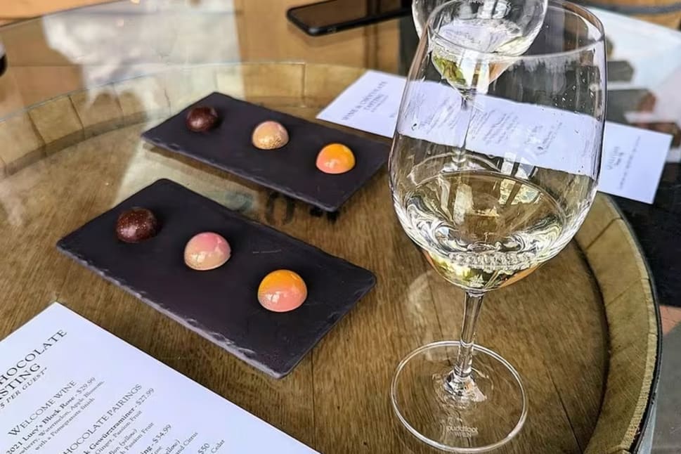 A glass of white wine and a menu placed on top of a wooden barrel, with chocolates and another glass of wine in the background.