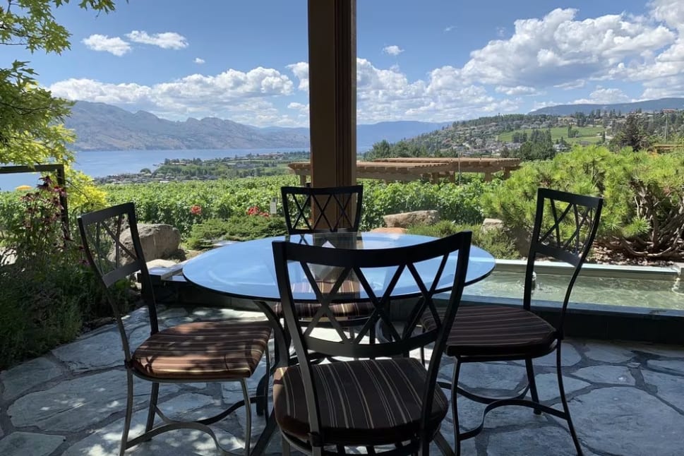a patio with a table and chairs and a view of a lake and mountains in the distance with a blue sky and white clouds