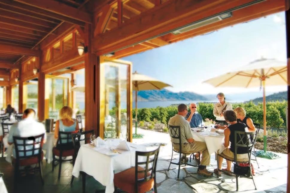 a group of people sitting at a table with umbrellas on the outside of a restaurant with mountains in the background at Quails' Gate Winery in Kelowna.