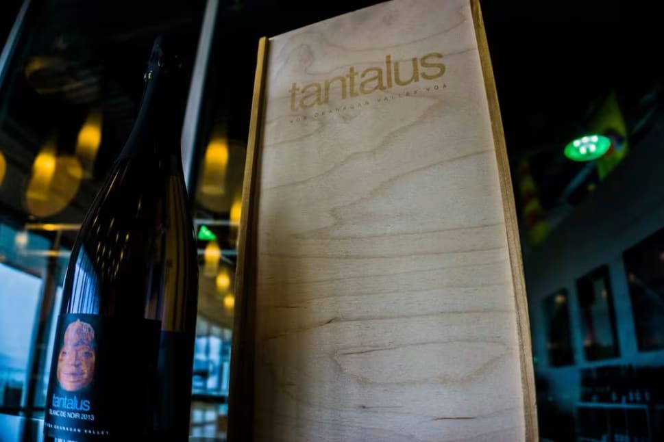 A bottle of wine sitting next to a wooden box on a table in front of a window with a sign that reads tantalus.