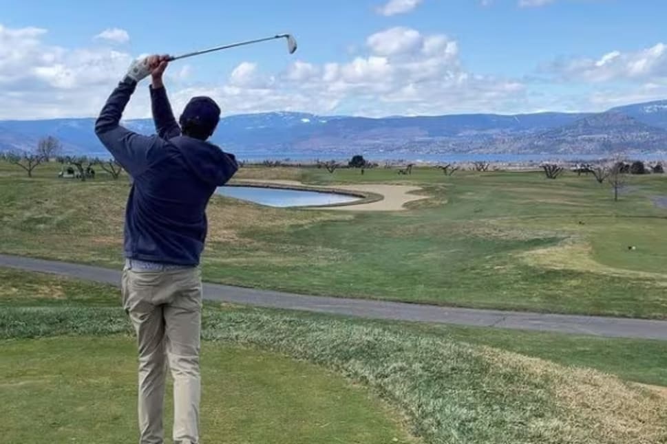a man hitting a golf ball with a golf club on a golf course near a lake and mountains in the distance at The Harvest Golf Club, Kelowna.