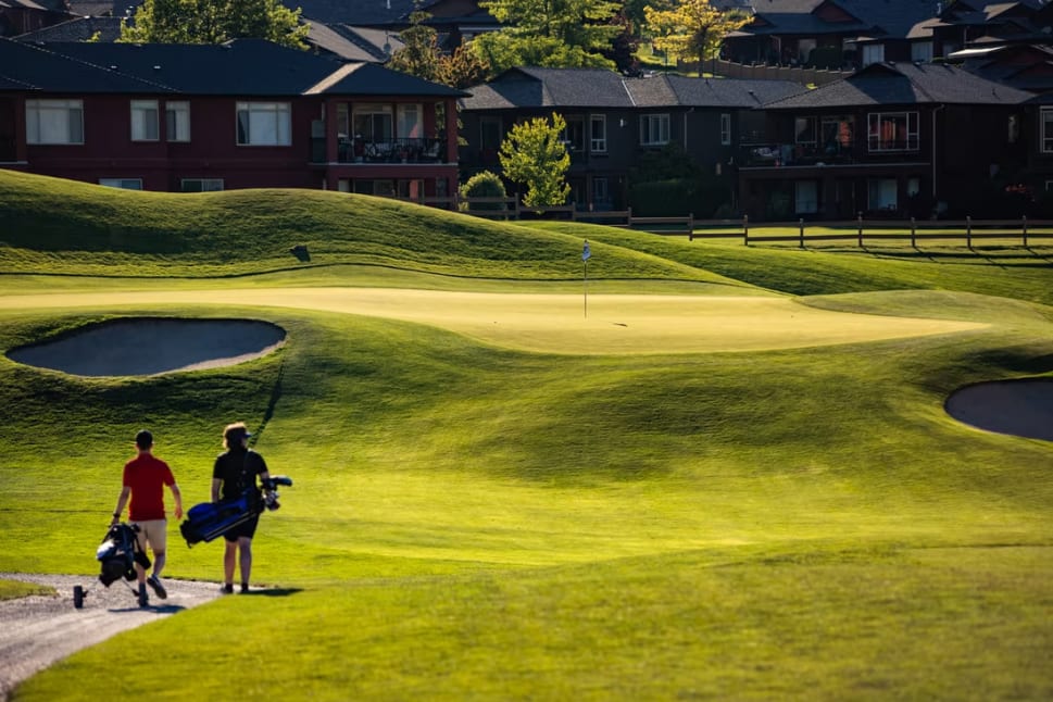 Two golfers walking towards a green on a sunny day at Two Eagles Golf Course in West Kelowna, with homes in the background.