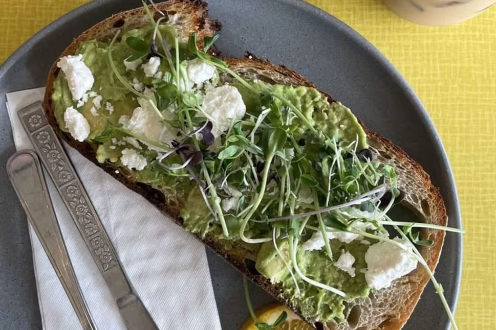 A slice of avocado toast topped with goat cheese and sprouts, served with a lemon slice on a grey plate along with a fork and knife, a glass of water, and an iced coffee on a yellow table at Sprout Bread in Kelowna.