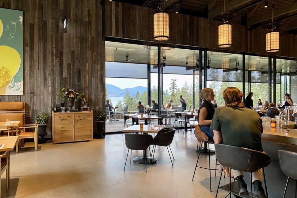 Spacious interior of Home Block at CedarCreek Estate Winery in Kelowna, with a cozy and rustic ambiance featuring wooden walls, large windows showcasing a scenic view, and patrons seated at tables and the bar.