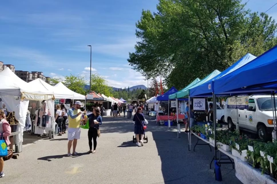 A group of people walking down a street next to tents with plants on them and a van parked on the side of the road at Kelowna Farmer's and Crafter's Market in Kelowna.