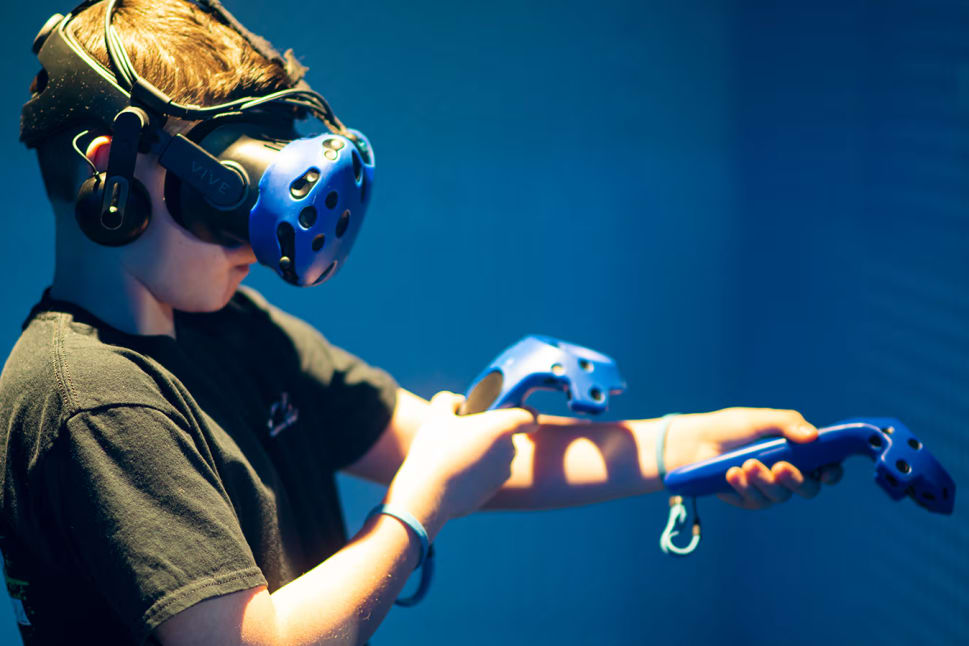 a boy in a black shirt is playing a game with a video game controller in his hand and a blue helmet on his head at Arcadia Virtual Reality Lounge in Kelowna