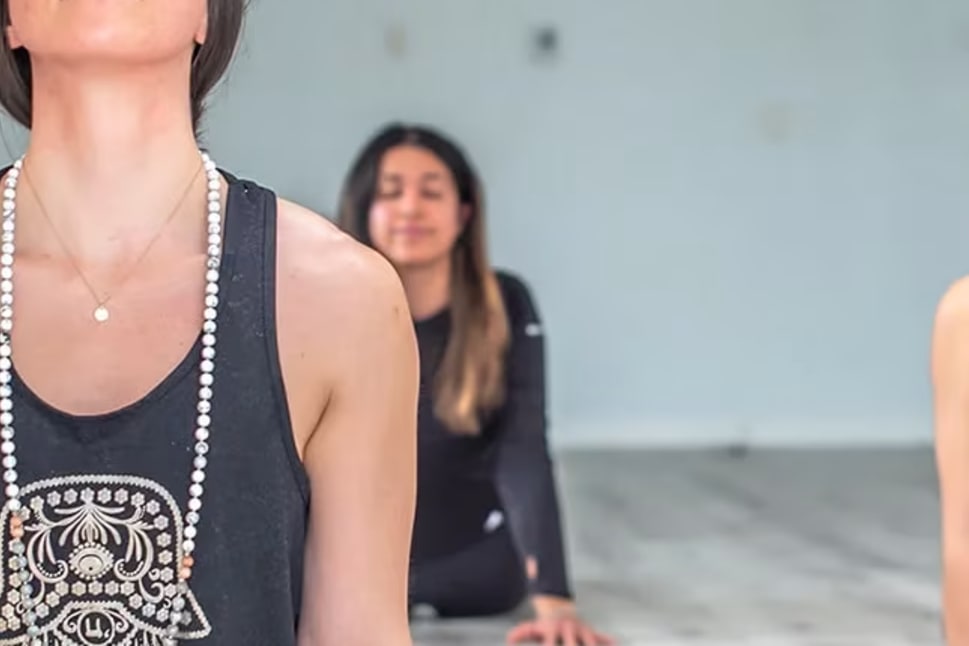 a group of women doing yoga poses in a room with white walls and wood flooring, one of the women is wearing a black tank top and the other is wearing a beaded necklace.