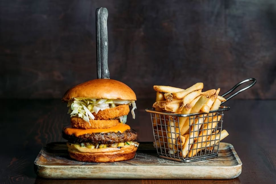 Loaded cheese burger with a kitchen knife holding it together. Side basket of french fries.