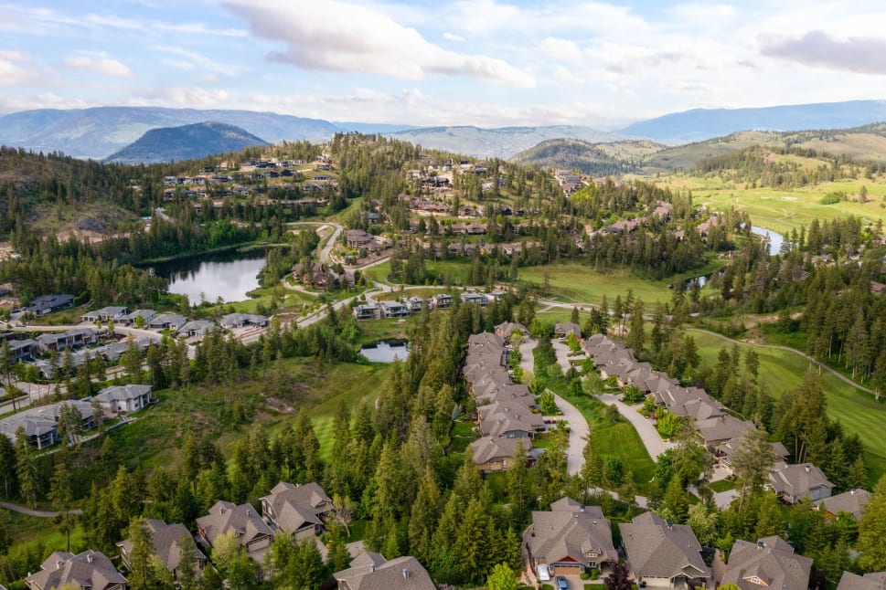 Aerial view of a residential area in Vernon surrounded by green hills, trees, and small bodies of water under a partly cloudy sky.