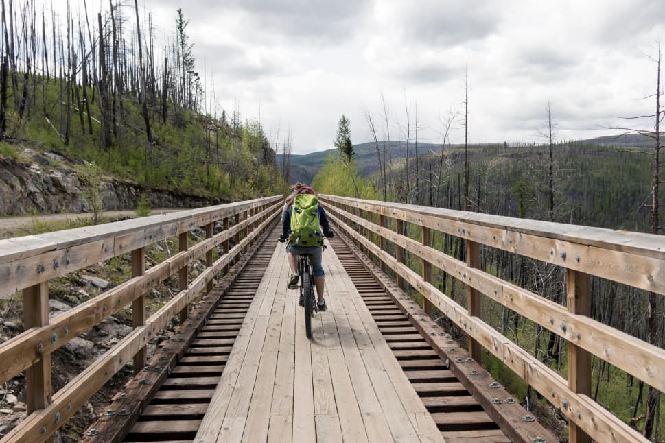A cyclist with a green backpack on a wooden bridge surrounded by lush greenery and a cloudy sky in Okanagan.