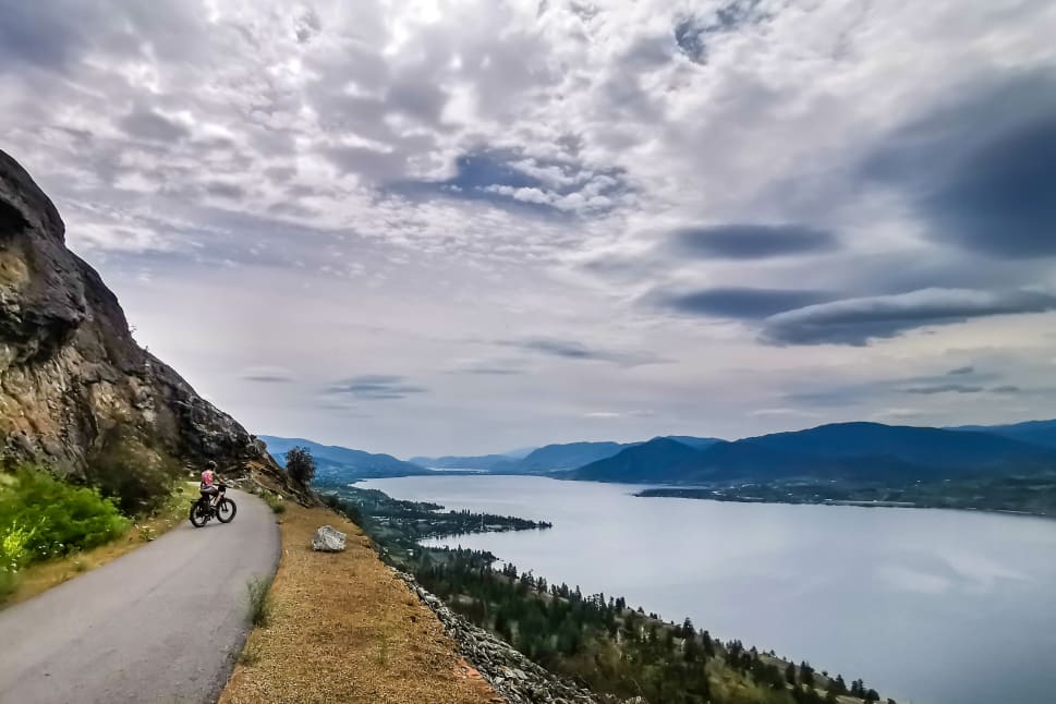 A cyclist riding along a scenic mountain path with views of Okanagan Lake under a cloudy sky.