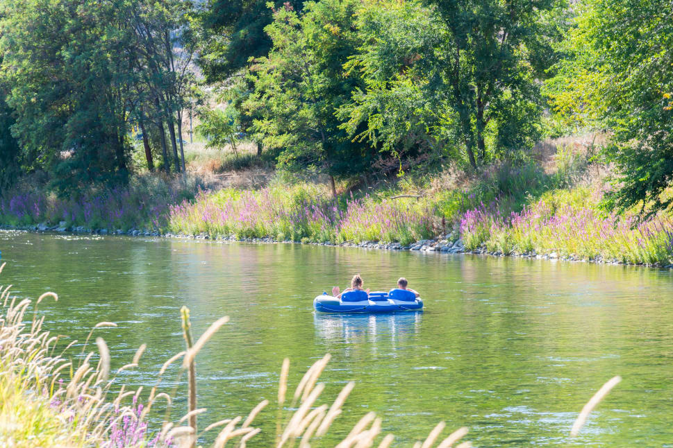People in a blue inflatable raft floating on a calm river surrounded by lush green trees and purple flowers in Penticton.
