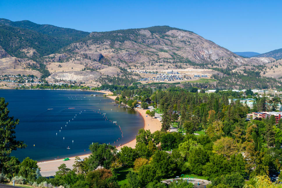 A scenic view of Penticton with a sandy beach along a blue lake, surrounded by greenery and mountains in the background.