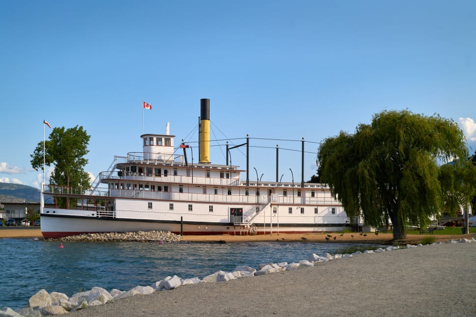 A historic paddlewheel steamboat docked on the shore in Penticton under a clear blue sky, with a tree and scenic background.
