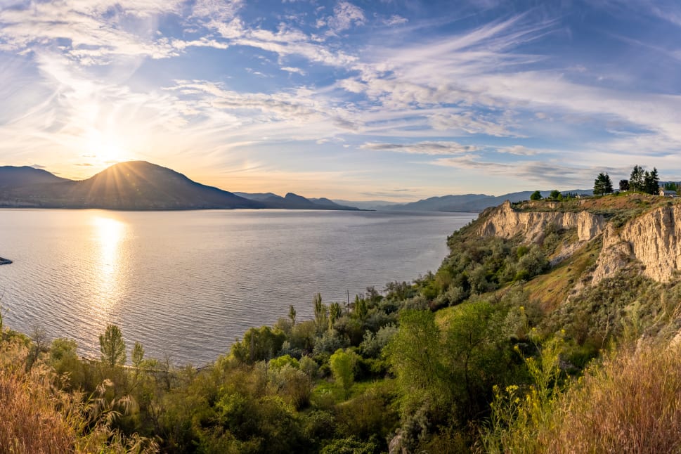 Panoramic view of a sunrise over Skaha Lake in Penticton with cliffs and vegetation in the foreground.