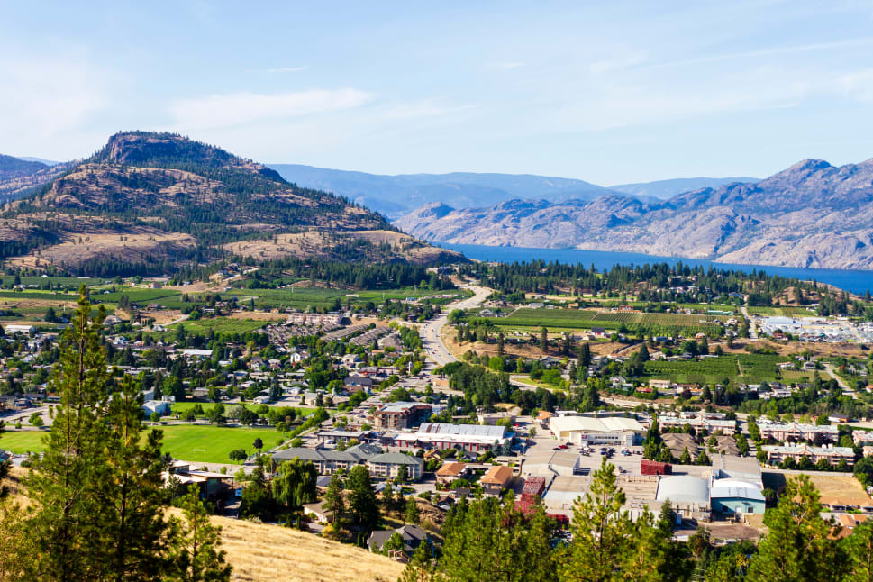 A panoramic view of Penticton, showcasing a mix of urban areas, green farmlands, and surrounding mountains with a lake in the background.
