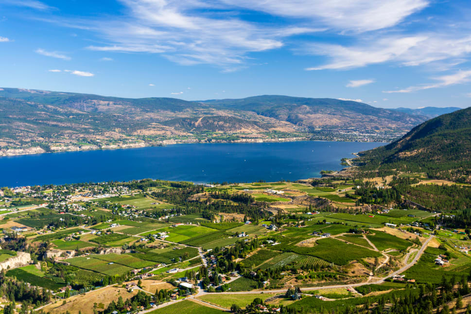 Aerial view of Penticton with a large blue lake, green agricultural fields, and surrounding mountains under a partly cloudy sky.