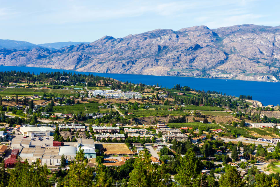 A scenic aerial view of Penticton with lush greenery, buildings, and a large lake bordered by rocky hills in the background.