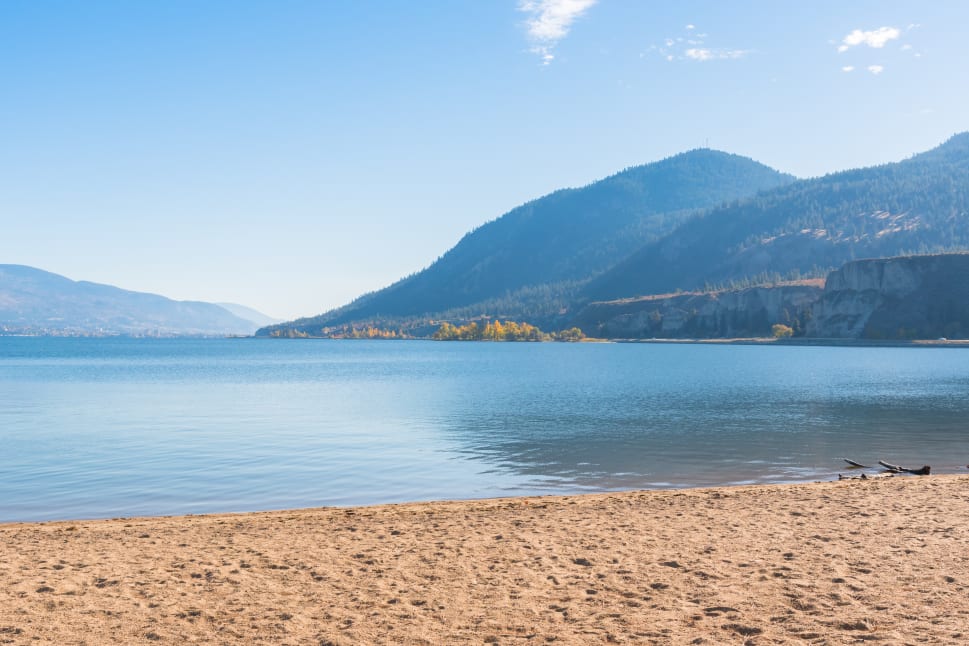 A tranquil view of a sandy beach leading to a calm lake with mountainous terrain in the background under a clear blue sky.