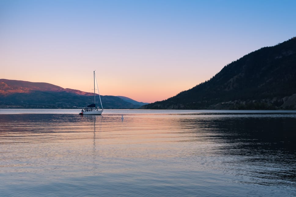 A sailboat rests on the calm waters of a tranquil lake at sunset in Summerland, surrounded by distant hills and mountains.