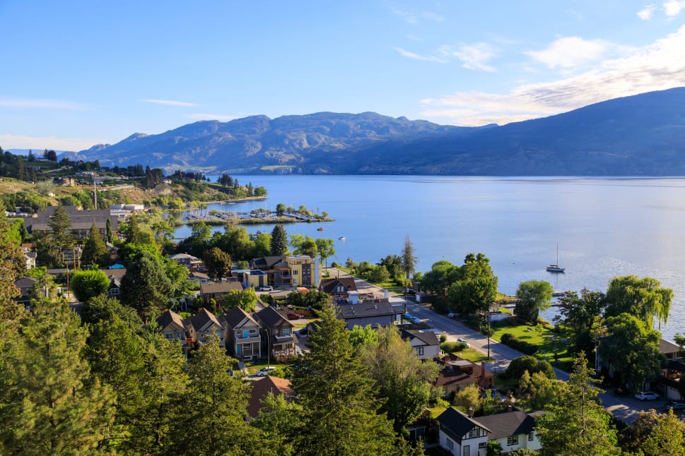 A picturesque view of Summerland featuring houses, trees, a marina, and a mountain range in the background by a serene lake.