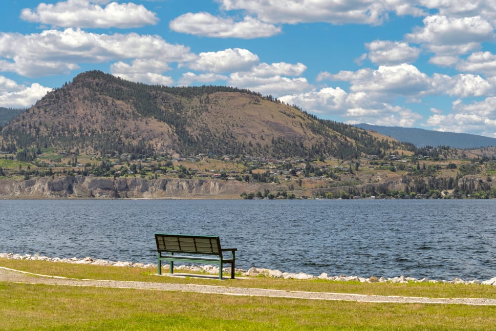 A bench overlooking a calm lake with a mountainous backdrop under a blue sky with scattered clouds in Peachland.